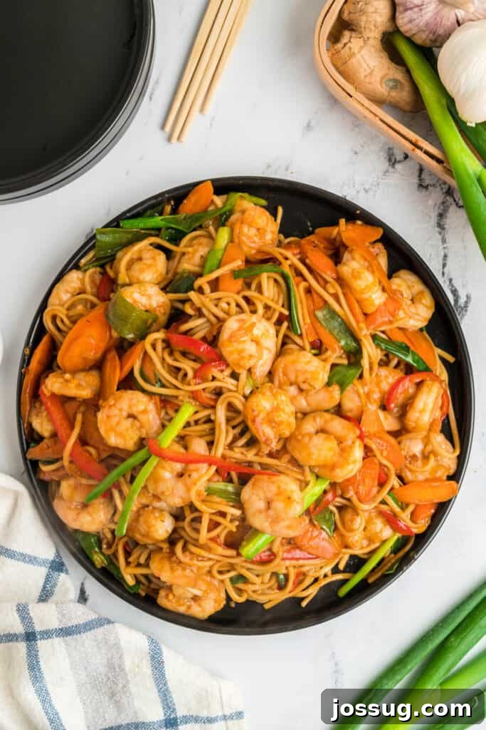Overhead view of shrimp lo mein on a black plate with shrimp, noodles, carrots, and green onions, garnished and ready to eat.