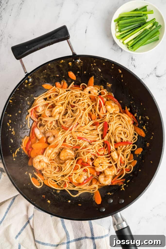 Shrimp, noodles, carrots, and bell peppers tossed together in a wok with sauce, illustrating the final mixing.
