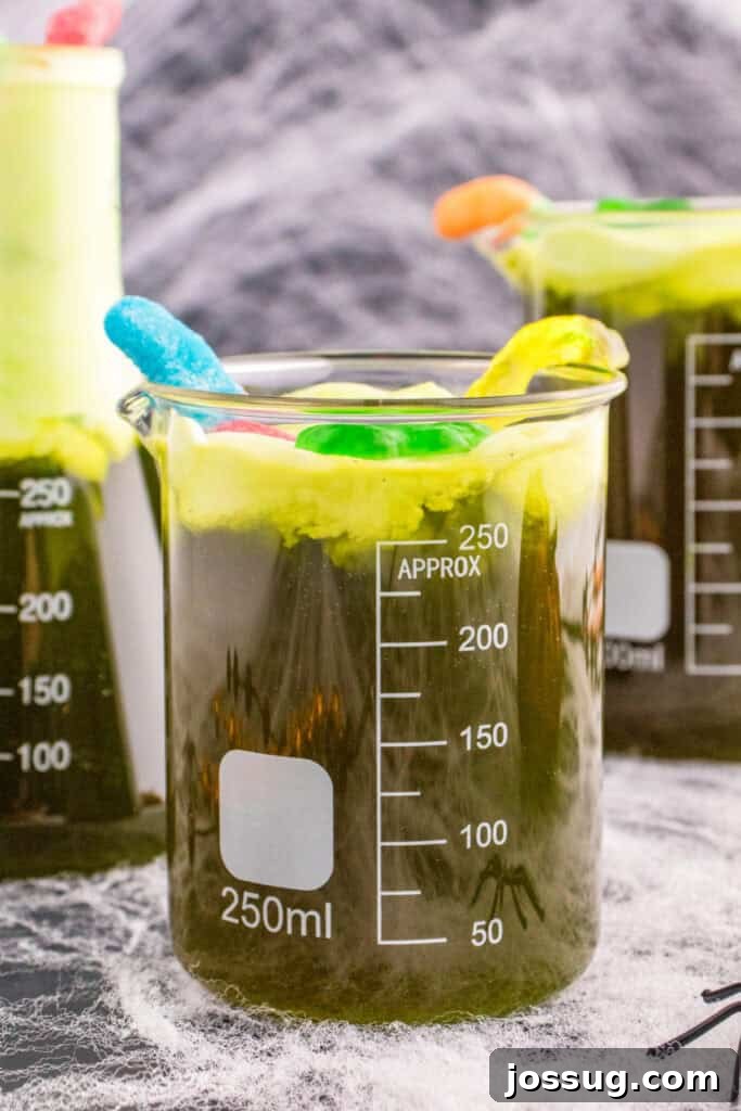 Biohazard Brew 2 Close-up of a beaker filled with bright green Toxic Waste Punch, topped with frothy green foam and an assortment of gummy worms and frogs, with cobwebs and a mysterious background.
