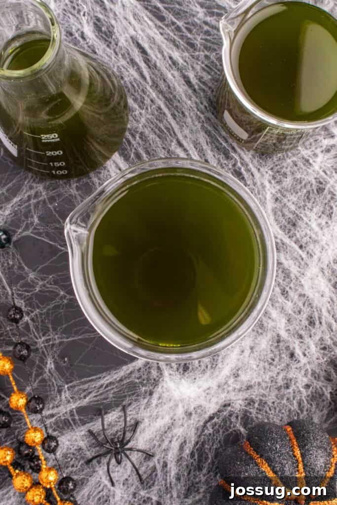 Biohazard Brew 14 Three glass beakers, filled to the brim with dark green Toxic Waste Punch, arranged on a cobweb-decorated table with scattered Halloween beads and miniature pumpkins for a themed display.