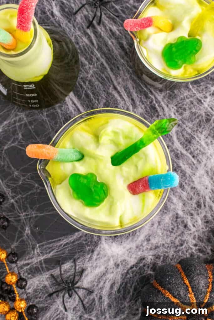Biohazard Brew 16 Beakers of Toxic Waste Punch elaborately decorated with playful gummy worms and vibrant gummy frogs on the foamy green topping, creating an utterly creepy and inviting Halloween drink.