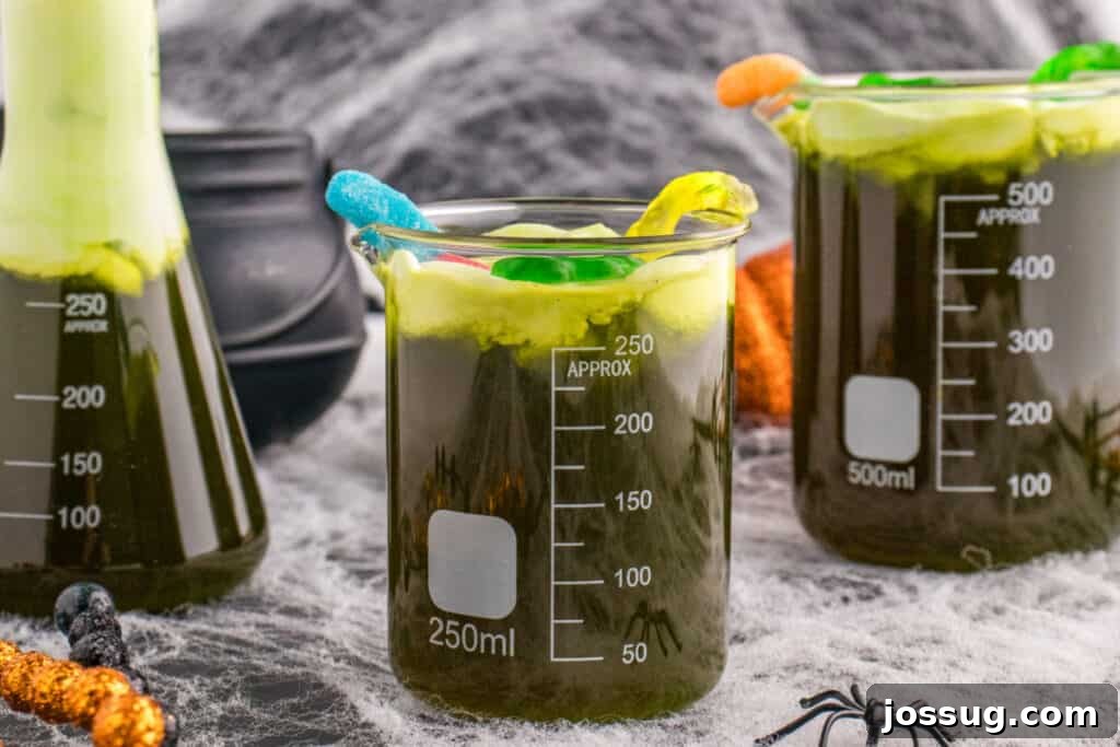 Biohazard Brew 18 A festive Halloween table adorned with multiple glass beakers of Toxic Waste Punch, each topped with a generous layer of green foam and whimsical gummy candy, all surrounded by intricate cobwebs and playful plastic spiders.