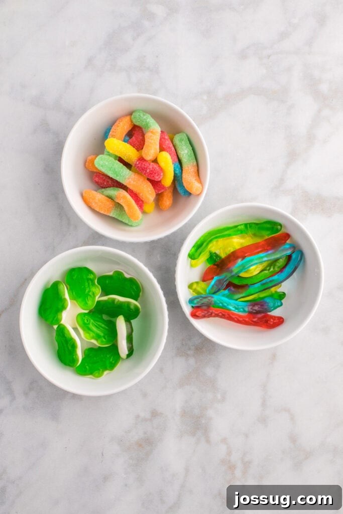 Biohazard Brew 5 Three small bowls brimming with colorful gummy candies, specifically sour worms, vibrant green gummy frogs, and slithering gummy snakes, ideal for punch decoration.