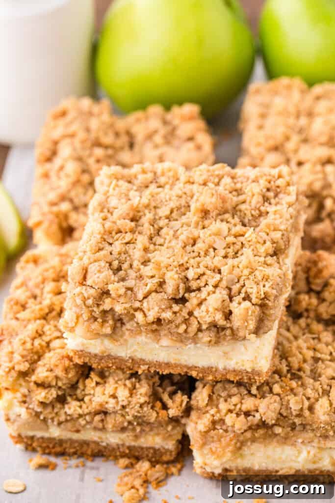 Stack of caramel apple cheesecake bars with visible apple filling and crumb topping, fresh apples in background.