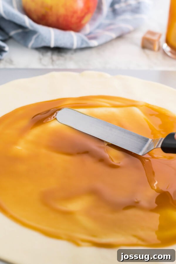Spreading thick caramel sauce evenly across the pie crust with an offset spatula, creating a rich, glossy layer.