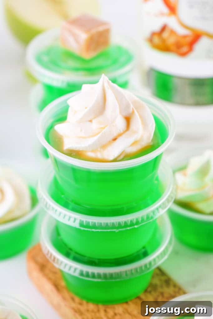 Stacked green caramel apple jello shots in plastic cups, topped with whipped cream and caramel drizzle.