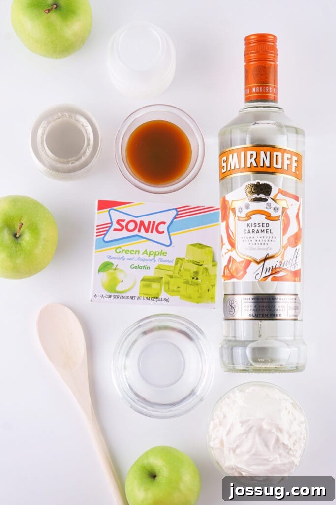 Ingredients for caramel apple jello shots including vibrant green apples, a box of Sonic green apple gelatin, a bottle of caramel vodka, a jar of rich caramel sauce, clean water, and fluffy whipped topping ready for assembly.