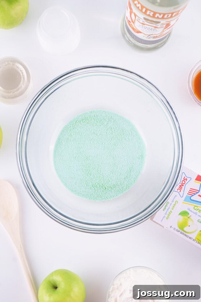Green apple jello powder poured into a clear mixing bowl, surrounded by ingredients for jello shots.