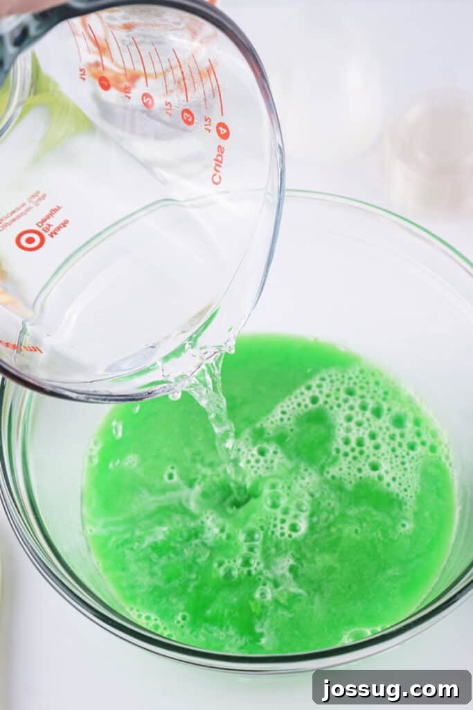Measuring cup of boiling water being poured into a bowl of green apple jello powder, creating a bright green mixture.