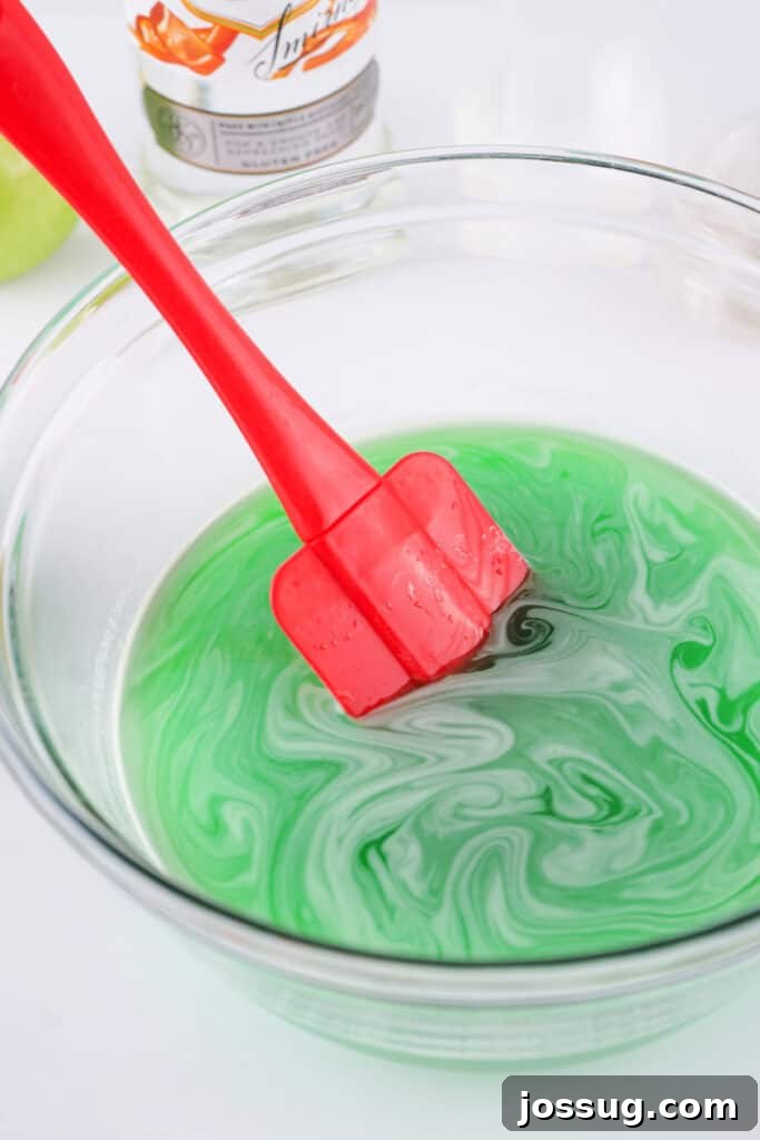 Red spatula stirring the dissolved green apple jello mixture in a clear glass bowl.