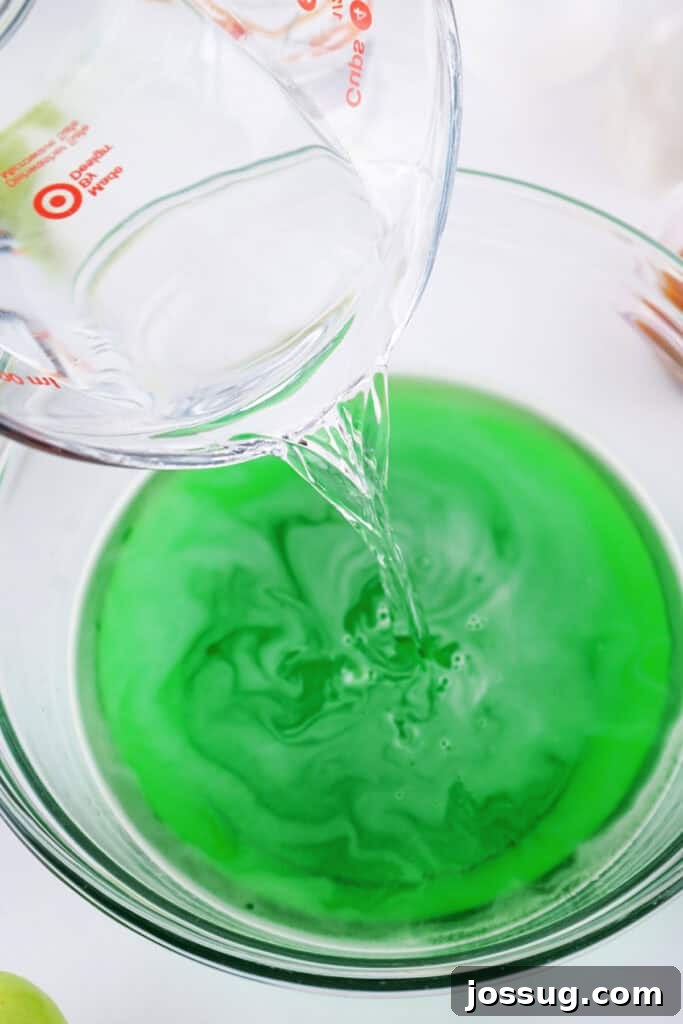 Measuring cup pouring vodka into the prepared green apple jello mixture.
