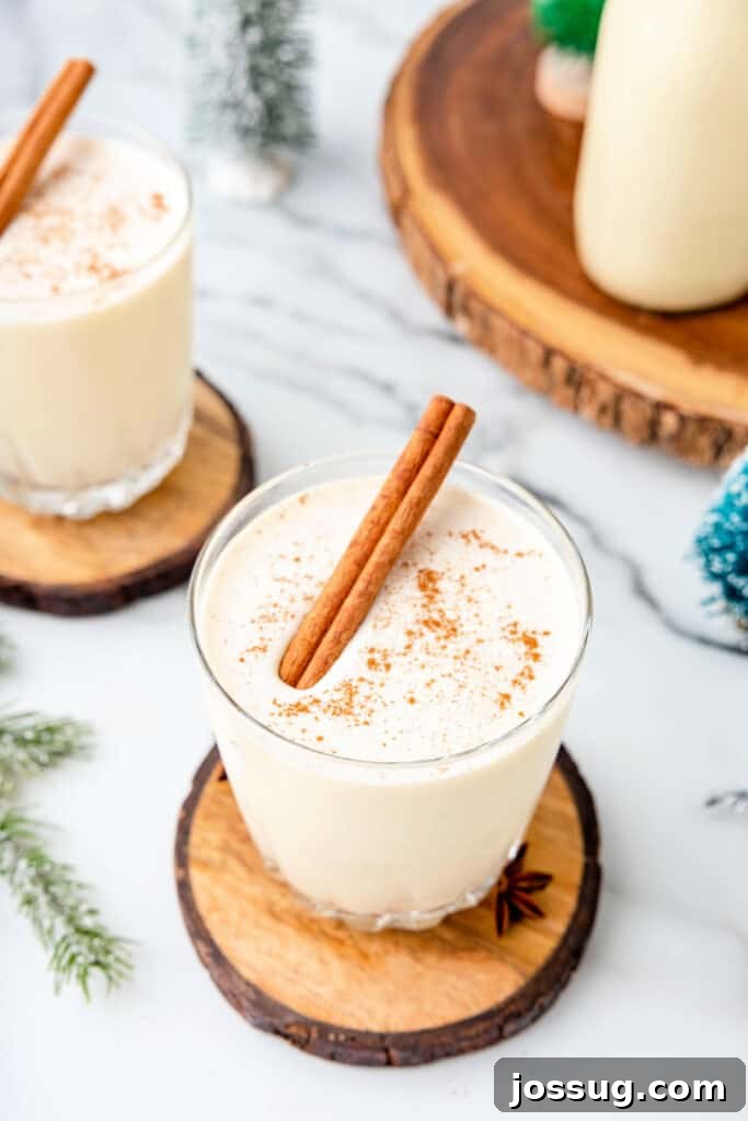 Classic Homemade Eggnog 2 Glass of homemade eggnog on a wood coaster, topped with ground nutmeg and a cinnamon stick, with holiday décor in background.