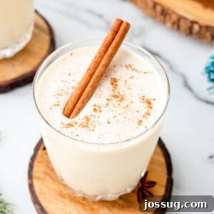 Overhead view of a glass of eggnog garnished with a cinnamon stick and nutmeg, styled with wood coaster and holiday greenery.