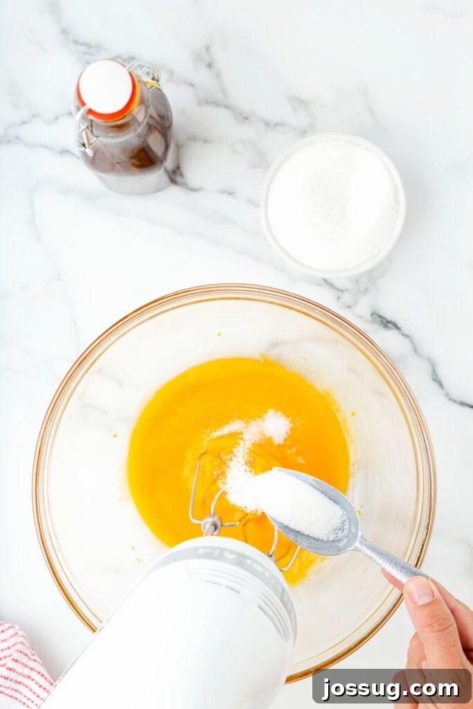 Classic Homemade Eggnog 5 Sugar being spooned into egg yolks in a glass mixing bowl with an electric mixer.