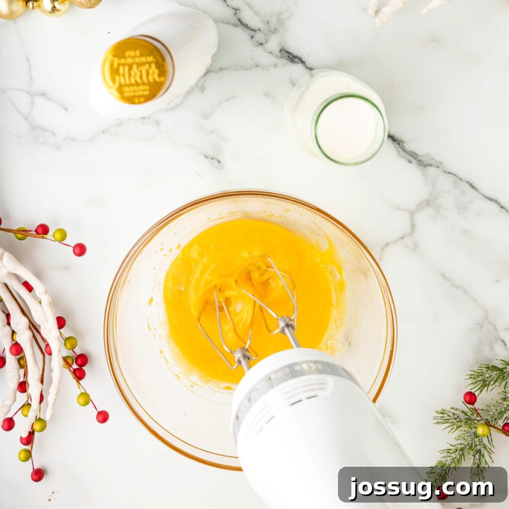 Electric mixer blending bright yellow egg yolks in a glass bowl on a decorated marble counter.