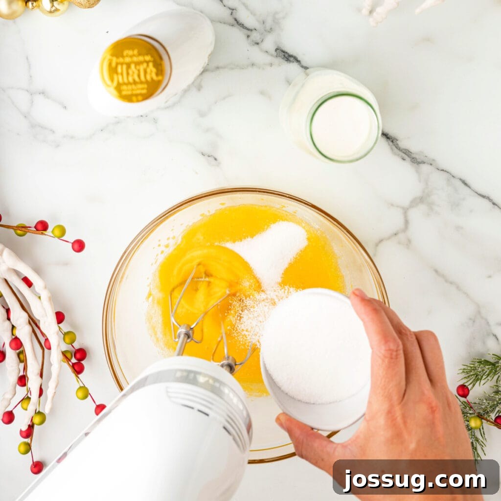 Hand pouring sugar into beaten egg yolks while using an electric mixer in a glass bowl.