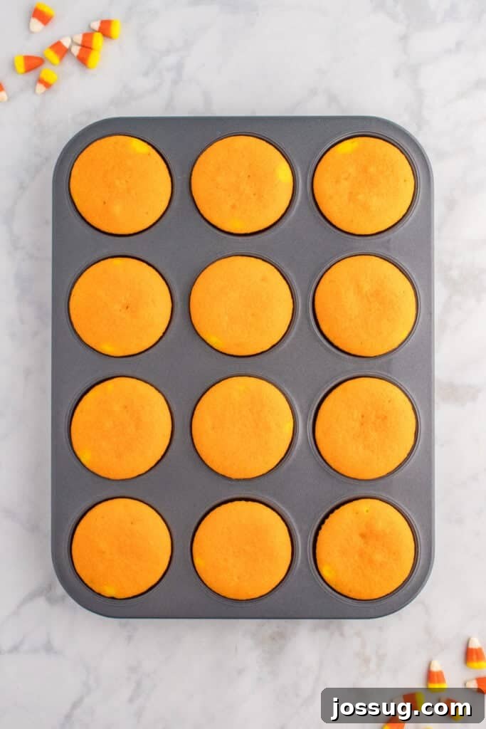 Baked orange cupcakes in a muffin pan cooling on marble surface with candy corn scattered nearby.