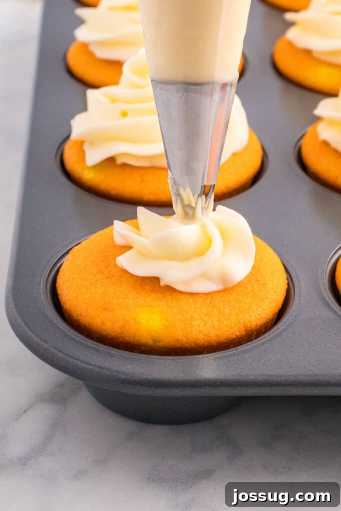 Piping bag swirling white frosting onto cooled orange cupcakes in a muffin pan.