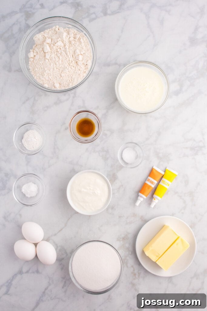 Overhead view of measured cupcake ingredients including flour, sugar, butter, eggs, yogurt, buttermilk, and food coloring on a marble surface.