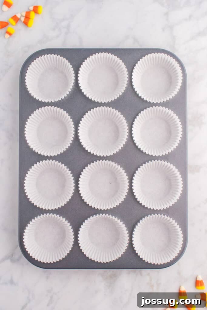 Muffin pan lined with white cupcake liners and candy corn scattered around on marble surface.