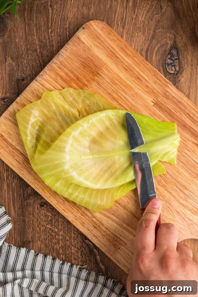 Hand using a knife to trim the thick stem from a softened cabbage leaf on a wooden cutting board.