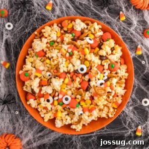 Square crop of Monster Mash Popcorn in an orange bowl with candy corn, candy pumpkins, pretzels, and eyeballs on a Halloween spiderweb background.