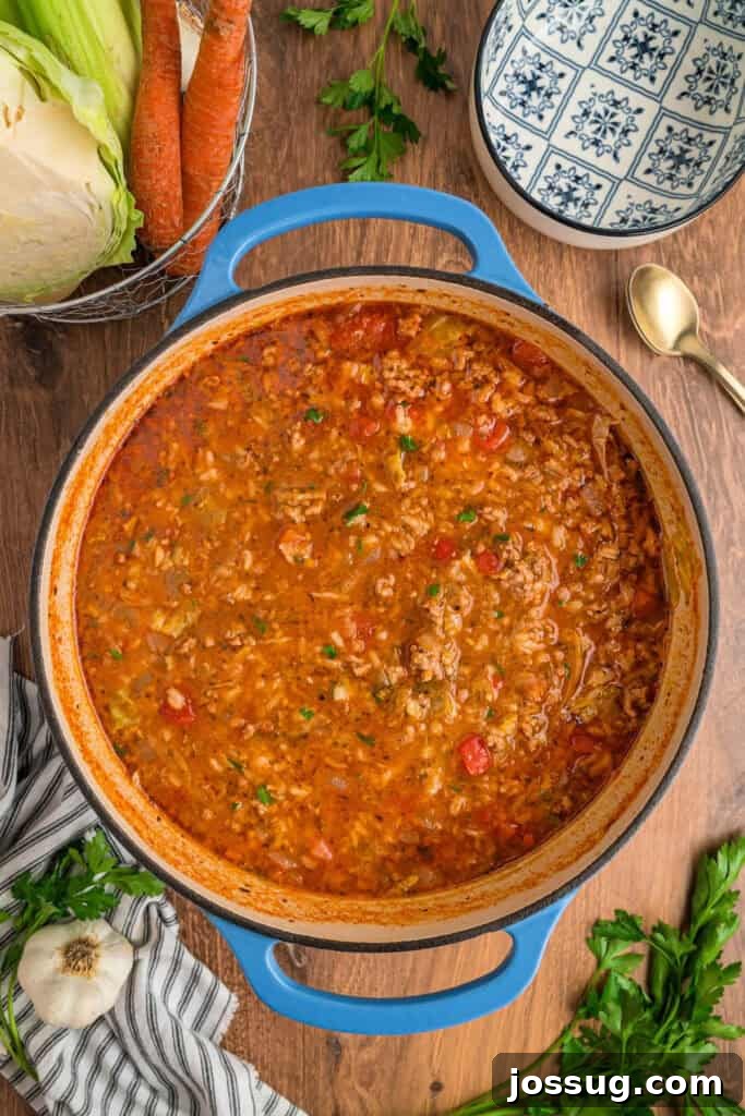 Finished cabbage roll soup simmering to perfection in a blue dutch oven, surrounded by fresh parsley, raw cabbage, and carrots on a wooden table.
