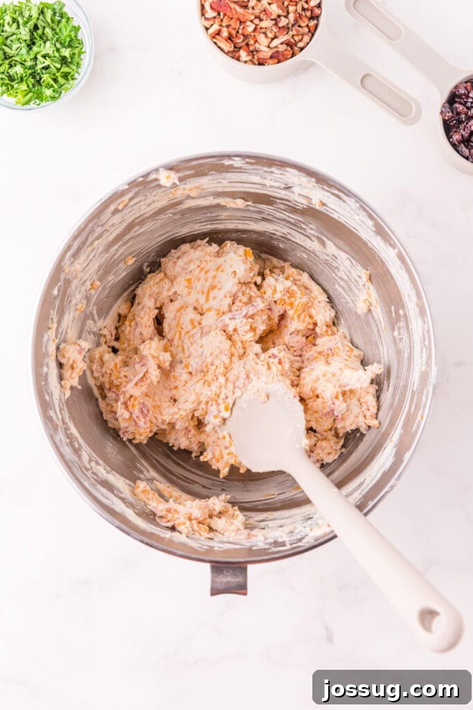 Mixed cheese and prosciutto filling in a stainless steel bowl with a spatula, showing texture of the Holiday Cheese Ball mixture.