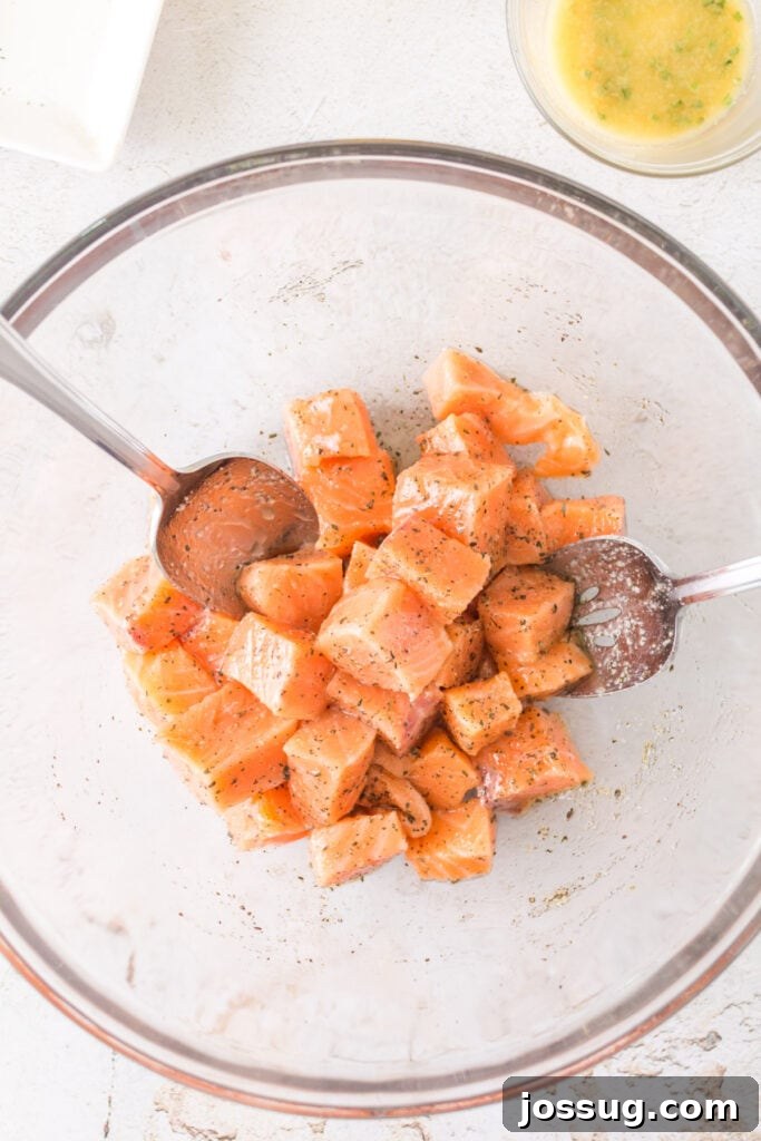 Cubes of raw salmon in a glass bowl being mixed with oil and seasonings using two metal spoons.