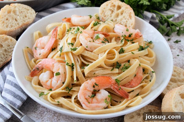 A white bowl filled with shrimp scampi pasta, garnished with parsley, with a slice of crusty bread on the side.