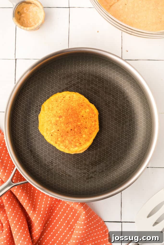 Golden pumpkin pancake cooking in skillet with visible bubbles and an orange towel beside the pan