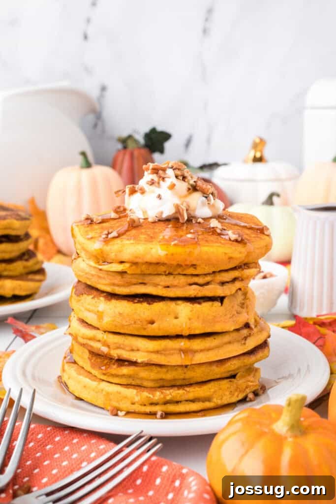 Tall stack of pumpkin pancakes topped with whipped cream, chopped pecans, and syrup surrounded by pumpkins and fall décor