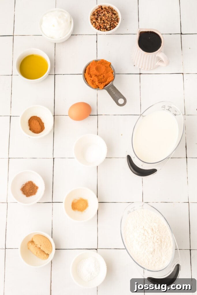 Overhead view of measured ingredients for pumpkin pancakes including pumpkin purée, milk, spices, flour, and egg on a white tiled surface