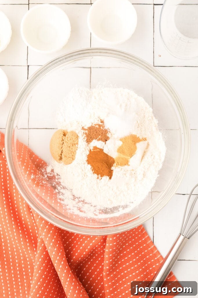 Mixing bowl filled with flour, brown sugar, and pumpkin spices on white tiled surface with orange kitchen towel