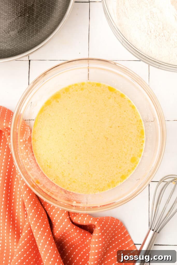 Whisked wet ingredients for pumpkin pancakes in a glass bowl beside a pan and orange towel