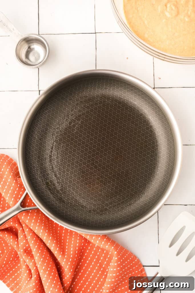 Oiled frying pan heating on stovetop beside pumpkin pancake batter and measuring cup