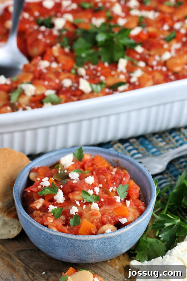 A small blue bowl holding a serving of Greek Baked Beans, topped with crumbled Feta cheese and fresh parsley, with the full casserole in the background