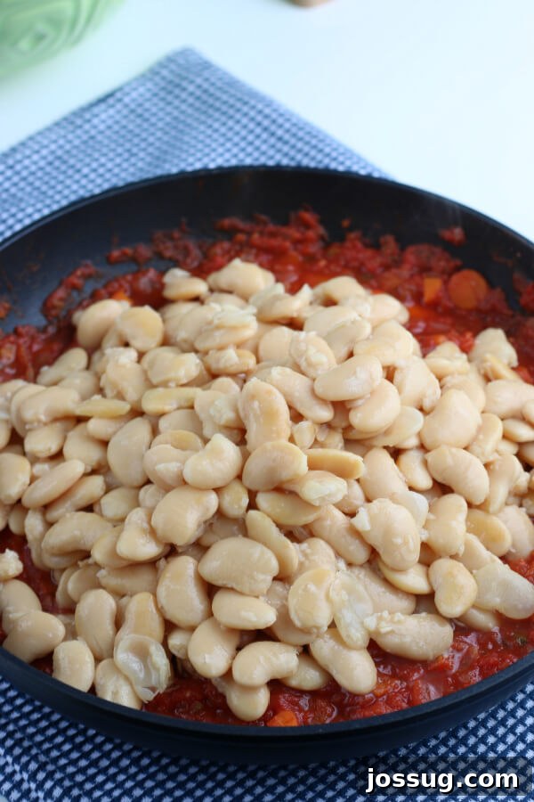 Cooked butter beans being added to the simmering tomato and herb sauce in a pan, blending the flavors
