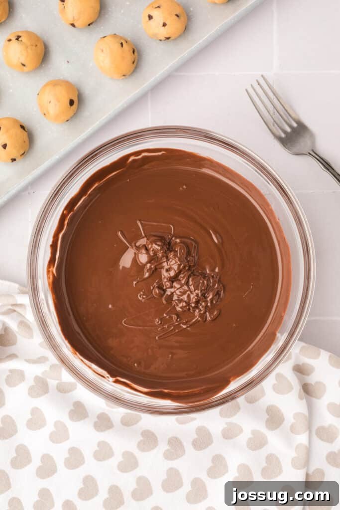 Bowl of glistening melted chocolate coating with a dipping fork resting beside it, and a baking sheet laden with chilled cookie dough balls ready for dipping.