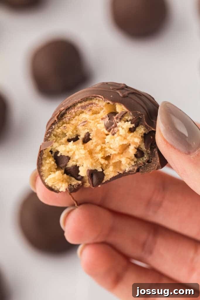 Hand elegantly holding a bitten chocolate truffle, revealing the perfectly formed edible cookie dough center studded with mini chocolate chips.
