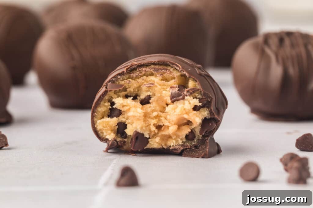 Close-up of a beautifully bitten chocolate-covered cookie dough truffle on a pristine white surface, with more delicious truffles softly blurred in the background, highlighting the creamy edible center.