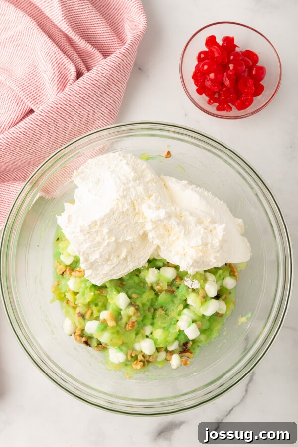 Green Jewel Salad 5 Thawed whipped topping being added to a bowl with the mixed ingredients for pistachio salad, alongside a small bowl of maraschino cherries.