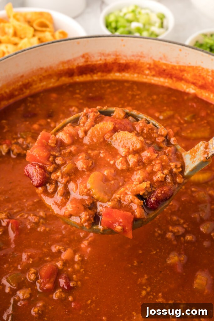 A serving ladle brimming with hearty homemade chili, just scooped from a large pot, showcasing its rich texture and vibrant ingredients.