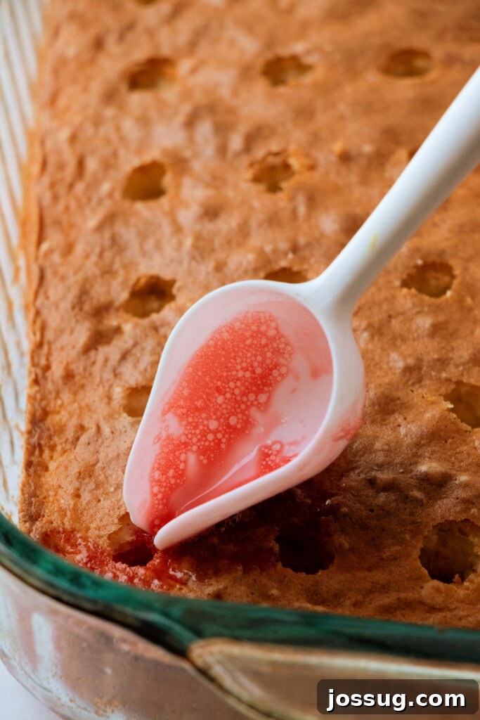 A hand carefully pouring red jello mixture into poked holes on a baked cake, illustrating the filling process