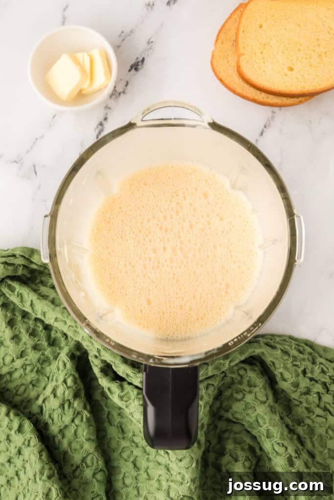 Eggnog Infused Brioche 5 Blended eggnog mixture smooth and frothy in blender bowl beside green towel and bread slices.