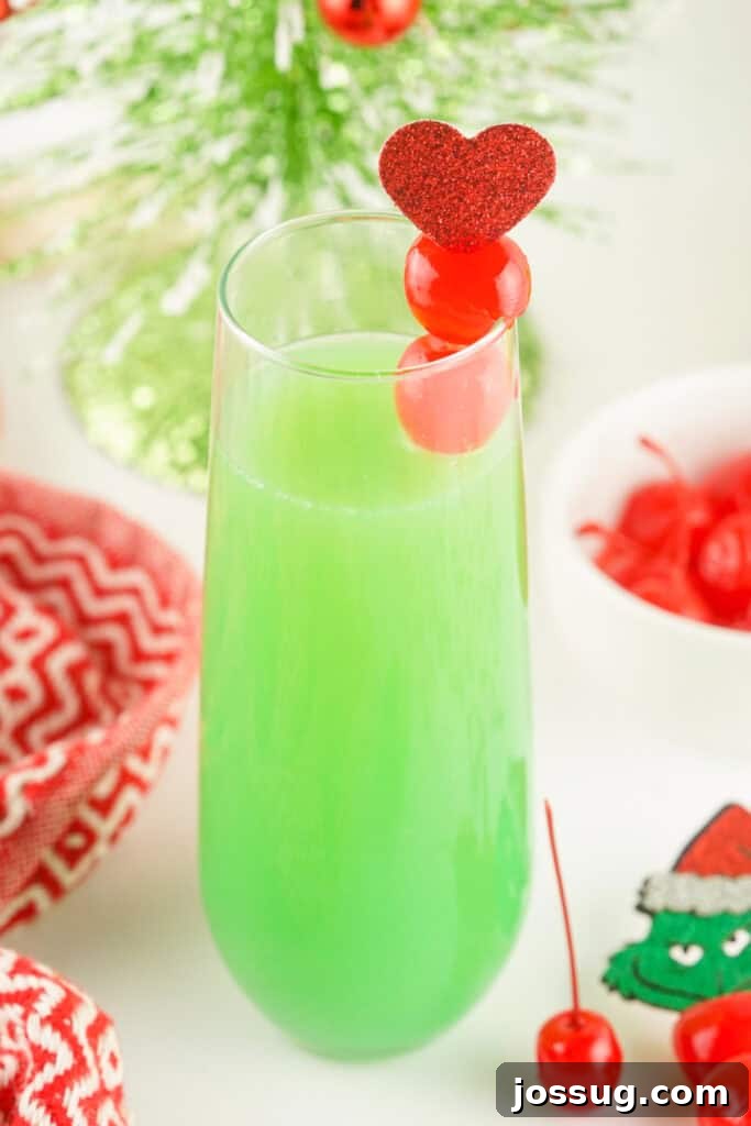 A vibrant green Grinch Mimosa in a champagne glass adorned with two maraschino cherries and a red glitter heart, capturing the essence of Christmas cheer.