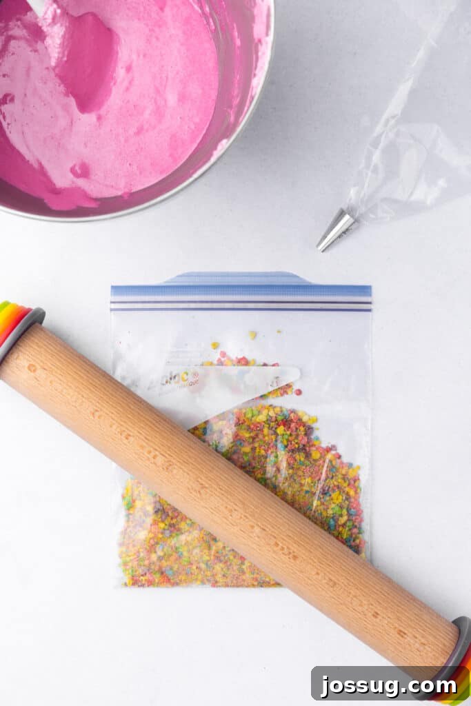 Pink macaron batter in a mixing bowl, piping bag fitted with a round tip, and crushed Fruity Pebbles in a zip top bag with a rolling pin