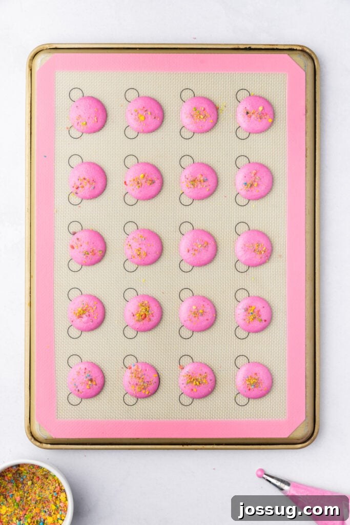 Pink macaron shells on a baking mat topped with crushed Fruity Pebbles before baking