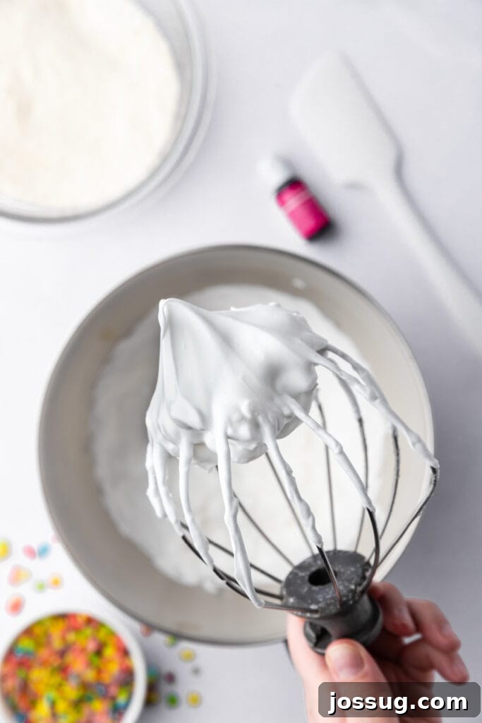 Fully whipped white meringue in a stand mixer bowl with gel food coloring nearby