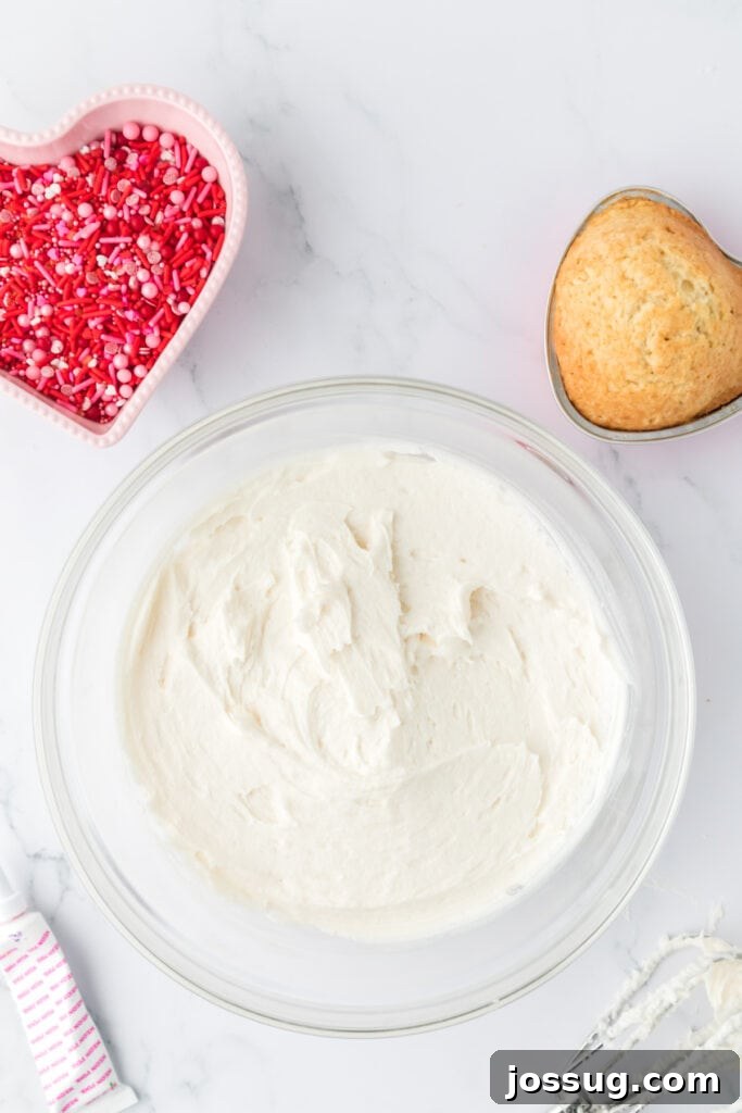Valentine's Day Mini Treat Cake 14 Large glass bowl filled with smooth white frosting, with a heart shaped pan of baked vanilla cake and a bowl of Valentine sprinkles nearby